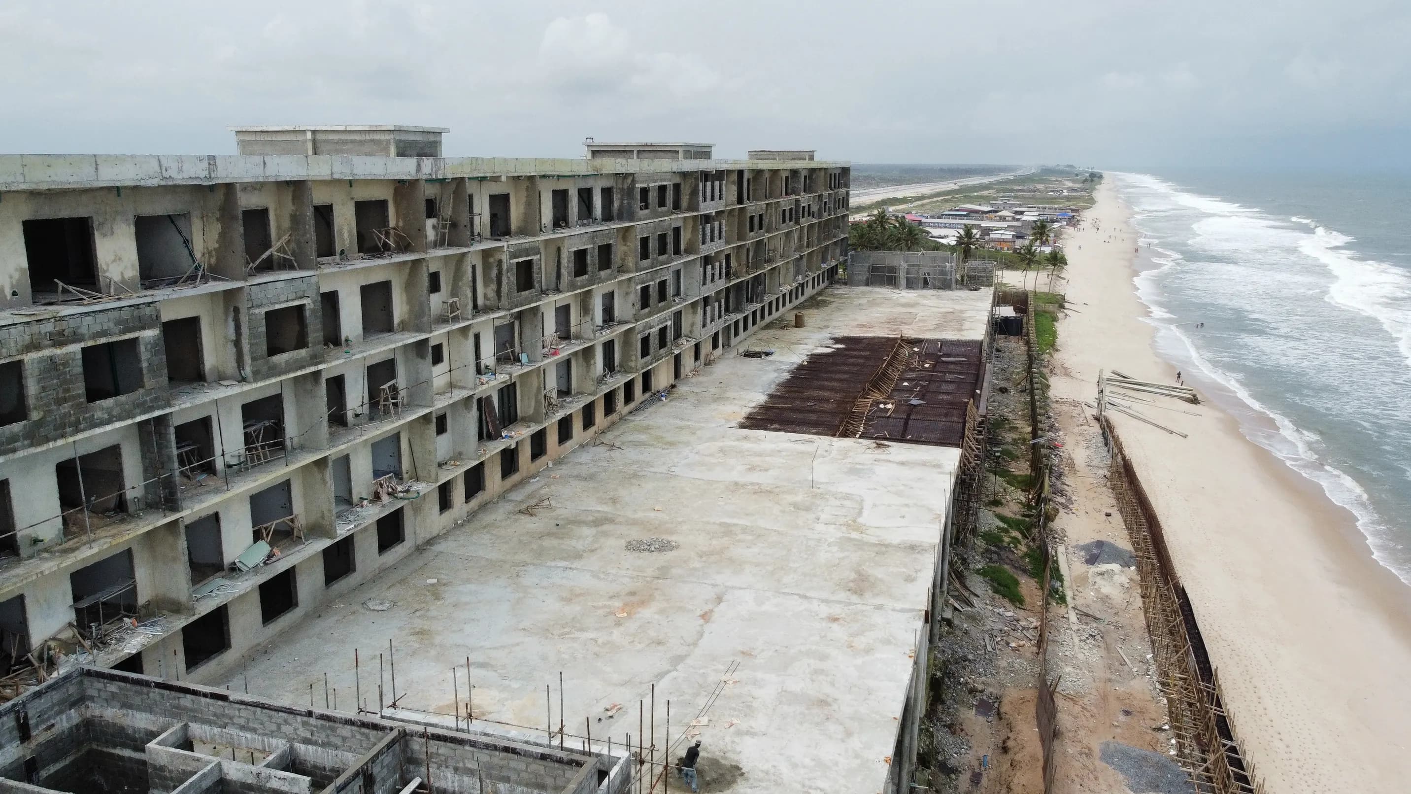 Aerial photograph of NJS Beach Resort under construction, showing the structural frame, exposed reinforcement starter bars, a cast podium slab, blockwork and the beach frontage beyond.